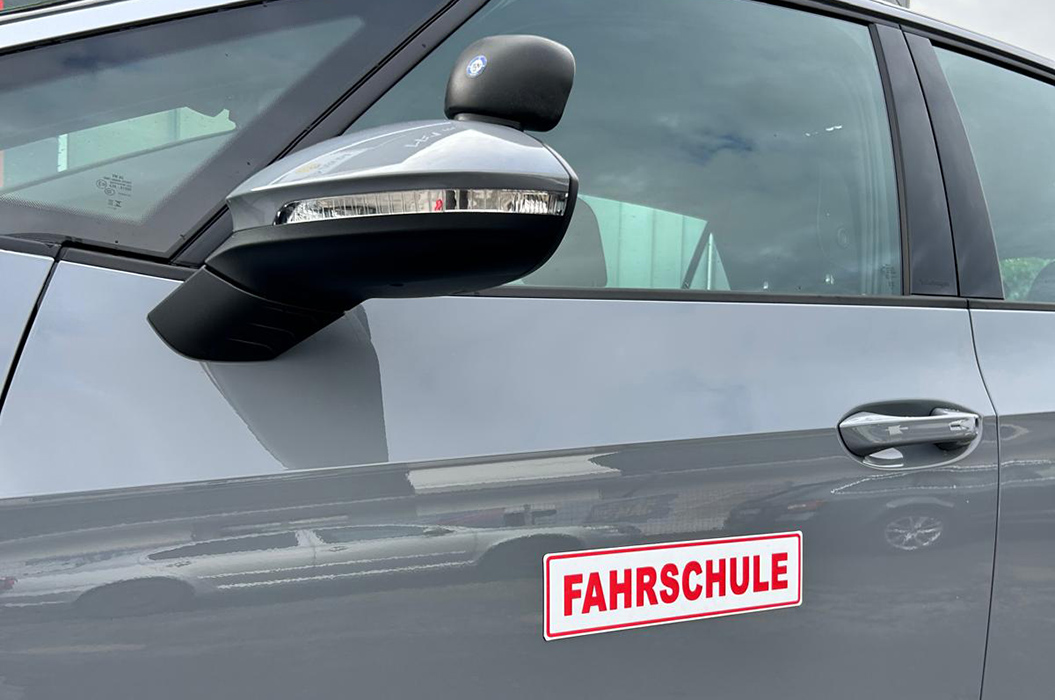 Graues Auto mit seitlichem Rückspiegel und rotem Fahrschule Schild an der Tür