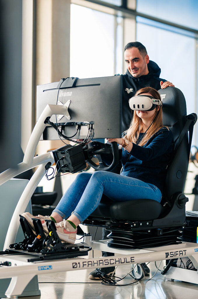 Eine junge Frau mit VR Brille sitzt konzentriert in einem Rennsimulator während ein Mann sie freundlich anlächelt